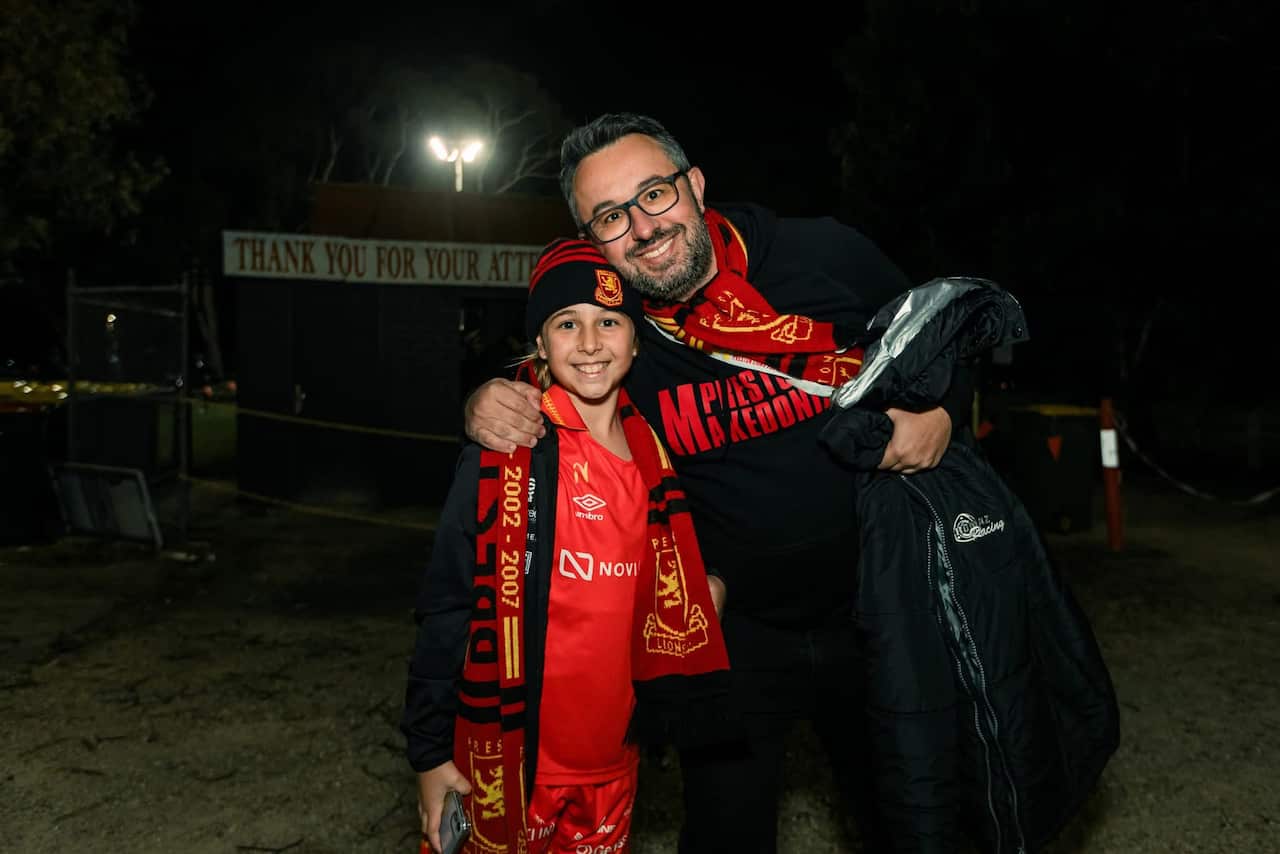 A man with dark hair and glasses wearing a red scarf smiles beside his daughter wearing the same scarf