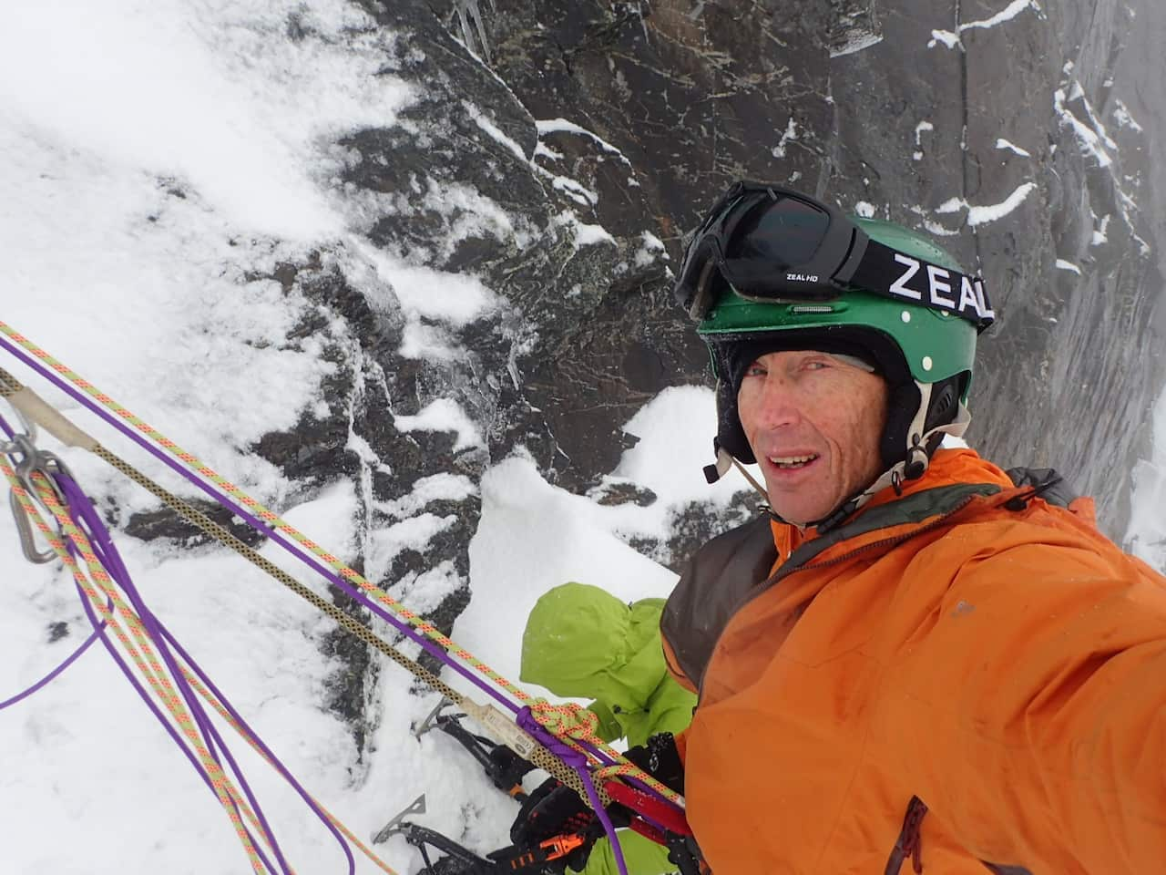 A man in an orange jacket clipped onto the side of a mountain by ropes.