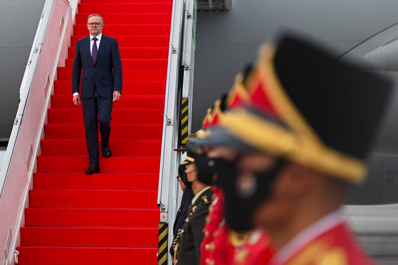 Australian Prime Minister Anthony Albanese walks down red stairs of the plane on arrival in Jakarta wearing a suit. Guards supervise his exit on the side. 