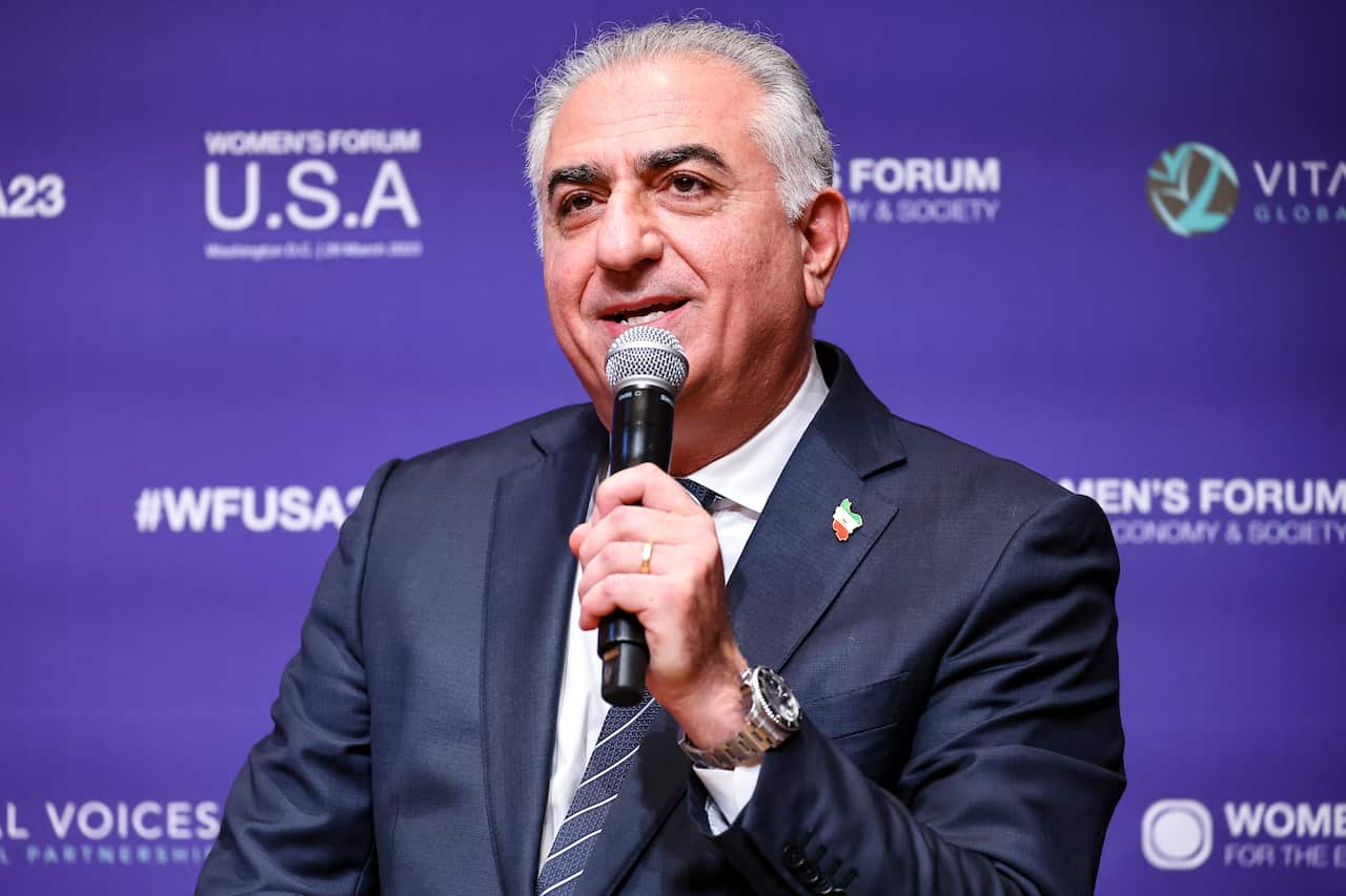 Reza Pahlavi, the exiled Crown Prince of Iran, speaking into a microphone at the Women's Forum USA event against a purple backdrop.