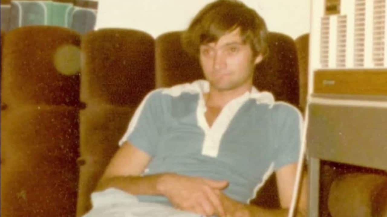 a vintage photo of a young man in a blue tshirt sitting on a brown velvet sofa