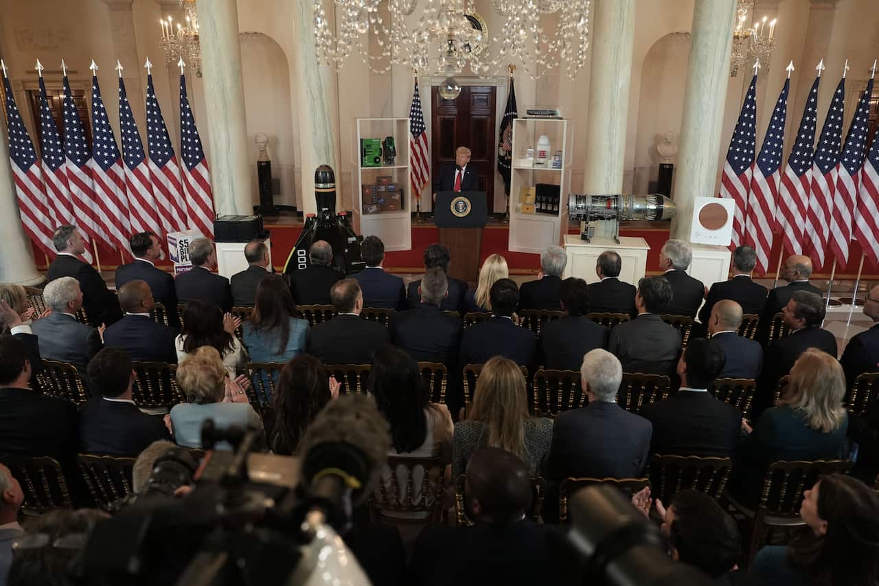 Seated crowd watch United States President, who is standing behind podium between American flags.