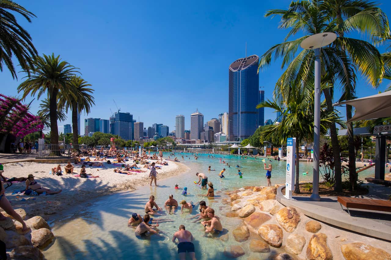People gather at a beach with white sand and palm trees. Many are sat in the shallow water, while some lie on the sand. In the background, there are high rise buildings.