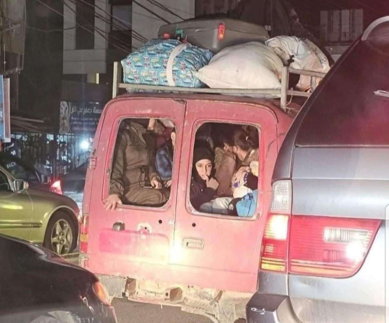 A pink van heavily loaded with luggage on its roof rack is filled with people looking out from the back windows onto a nighttime street crowded with traffic.