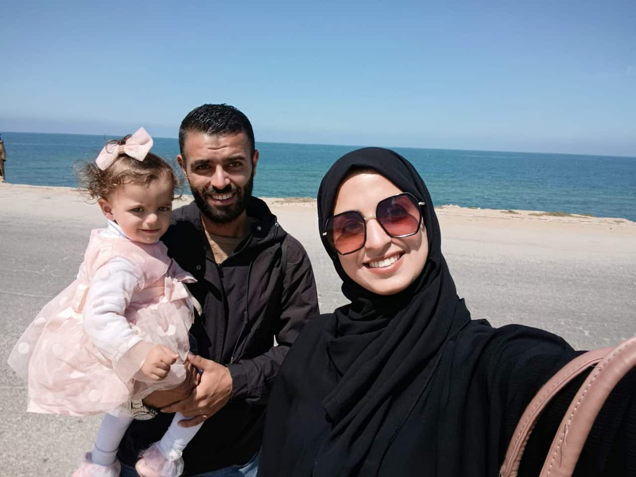 A woman wearing a black hijab takes a selfie with a man holding a small child in a pink frock. They are on a beach with the sea in the background