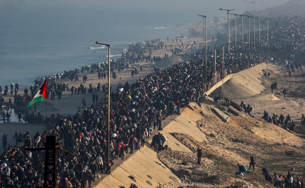 A massive crowd of people walk along a coastal road and sandy beach under a hazy sky.