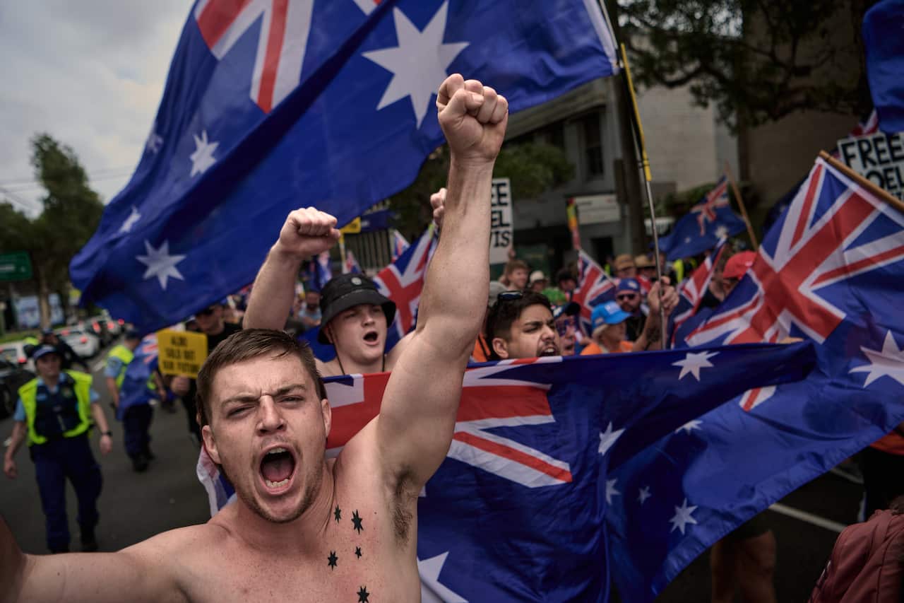 Un hombre sin camisa encabeza una multitud que porta banderas australianas. Parece estar gritando y tiene el puño en alto.