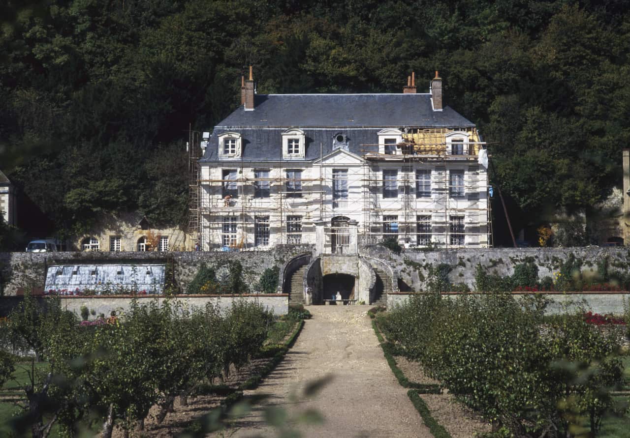A large stately home with scaffolding across its front. A long tree-lined driveway leads towards the house. 