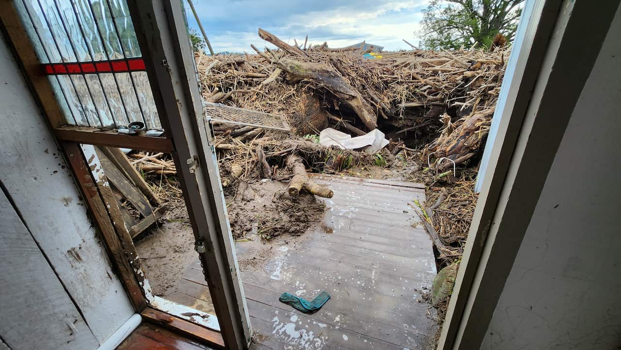A huge, dense wall of debris including branches and pieces of doors, pushed up against the back porch of a home. The photo taken from inside the home, shows the back door, smashed side window, and how close the debris has been pushed up against the house.