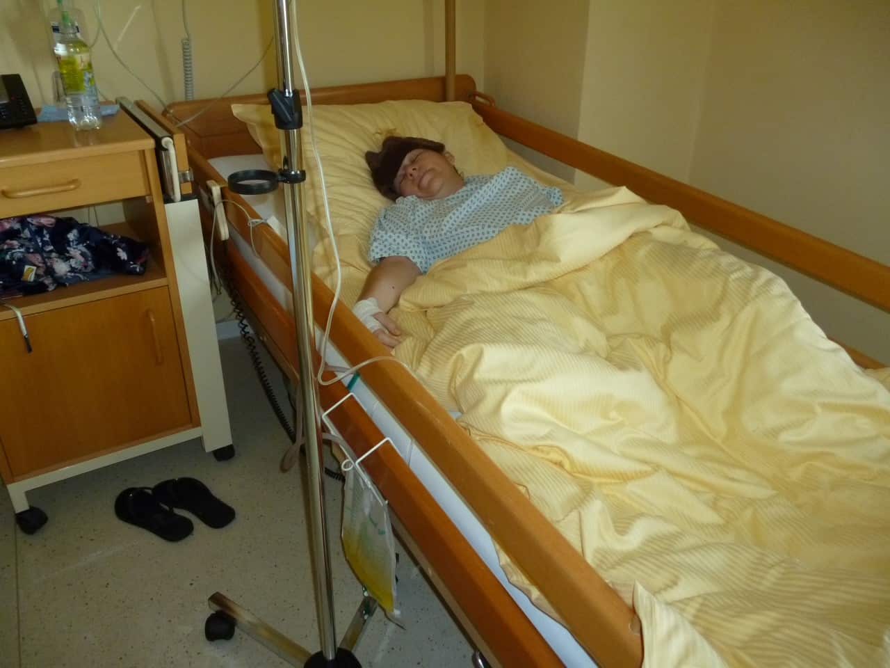 A woman lies asleep on a hospital bed under yellow blankets while receiving a drip.