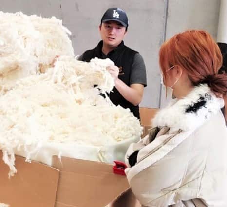 A man in a black vest stands next to a mound of wool, with another person in foreground.