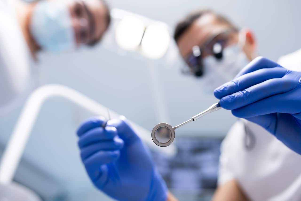 A dentist wearing blue disposable gloves and white scrubs looks down from above while holding a dental instrument.