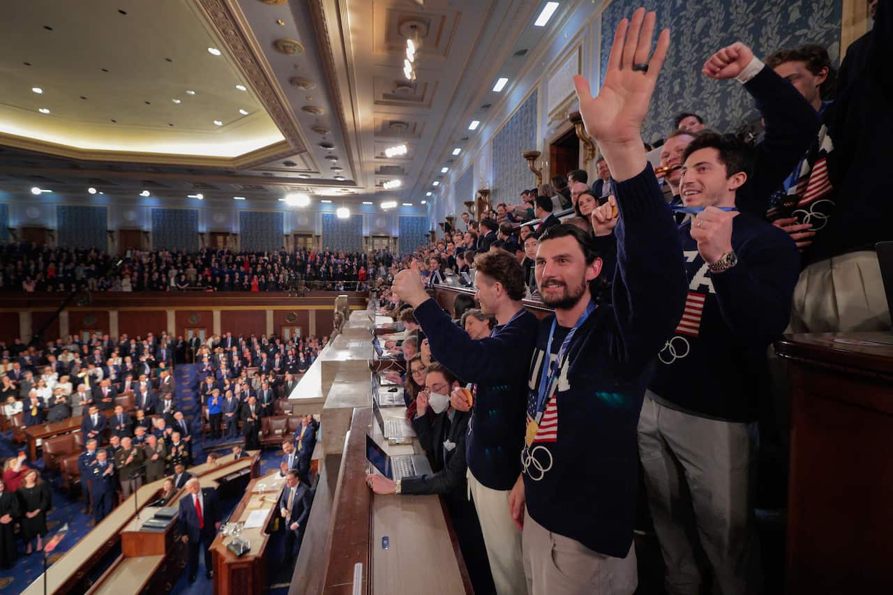 A group of men wearing tops with the American flag and wearing medals waves to a crowded room.