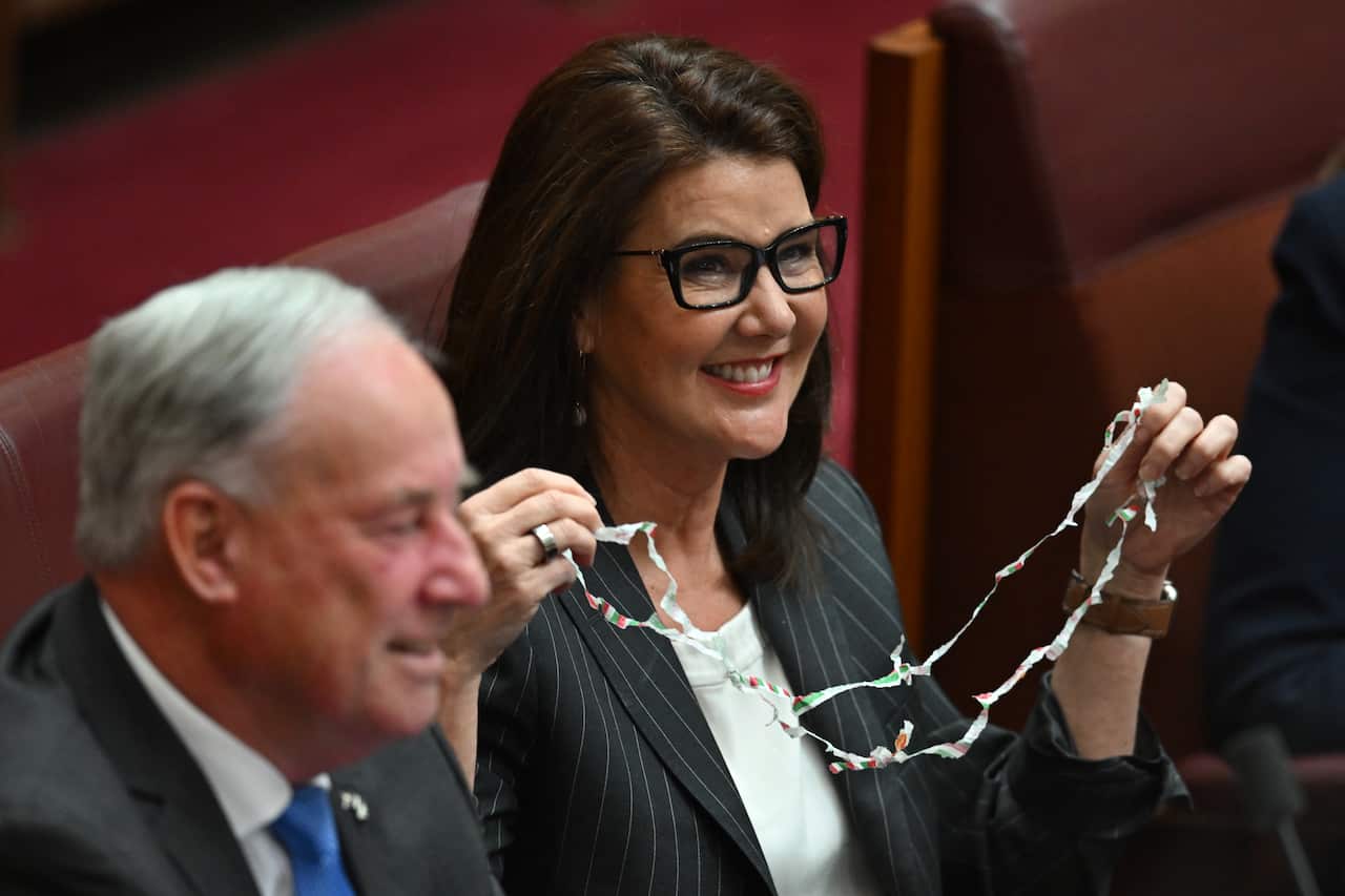 Senator Jane Hume, in thick black glasses, holds up a paper chain made up of mintie wrappers.