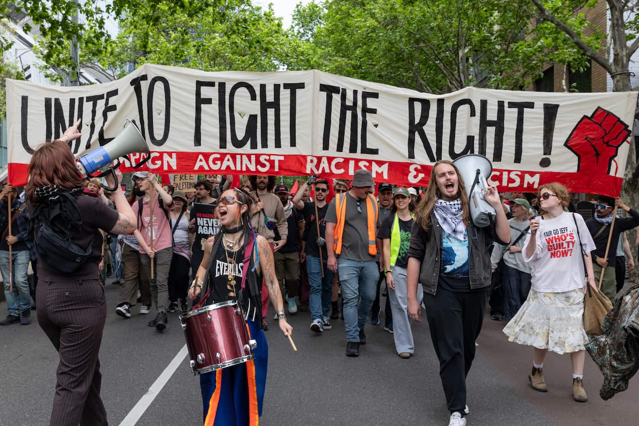 A group of protesters holding a banner during a rally, with some in the lead carrying drums and speakers.