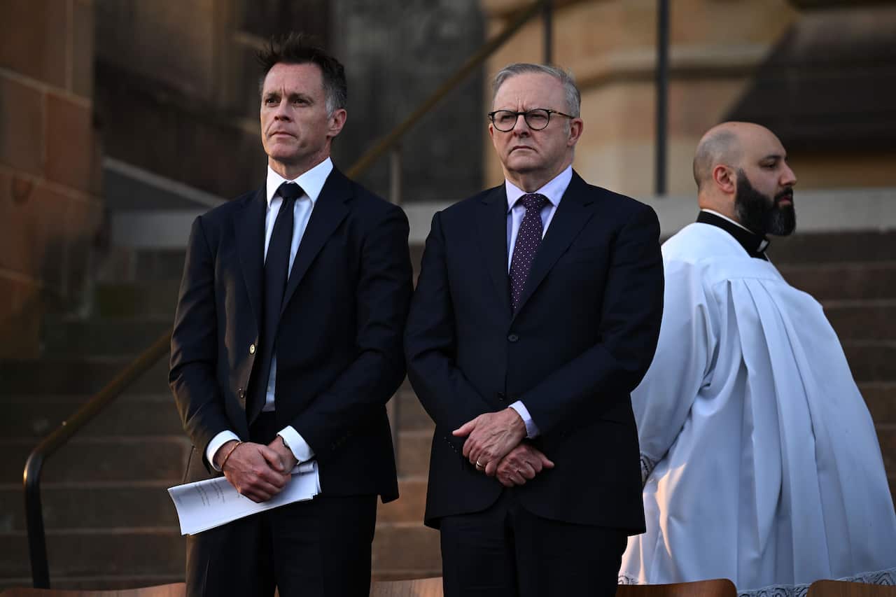 Two men in suits are standing with their hands clasped in front of them. There is a man in a white robe standing next to them.