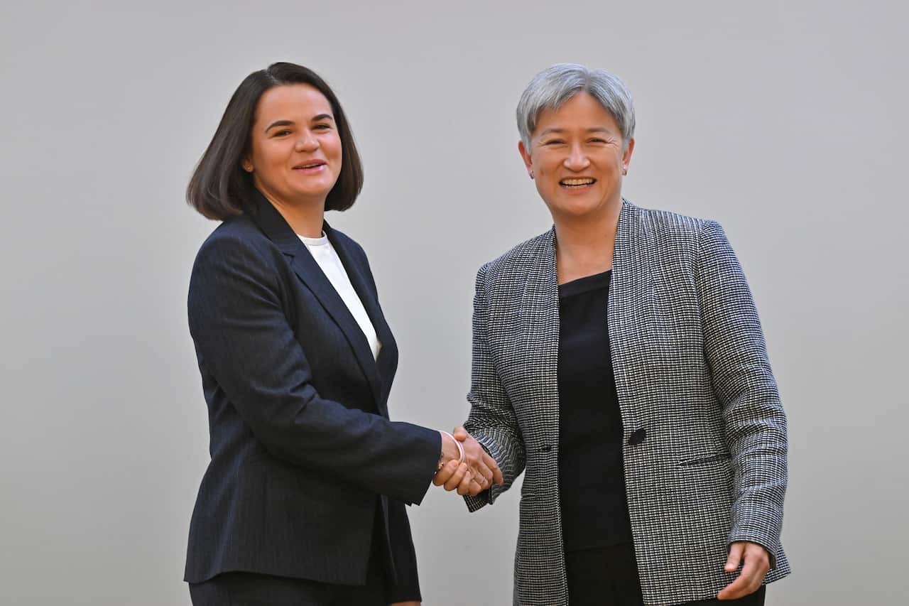 A woman on the left, wearing a dark suit and white top, shakes hands with Penny Wong on the right, wearing a grey-and-white patterned blazer. Both are smiling.