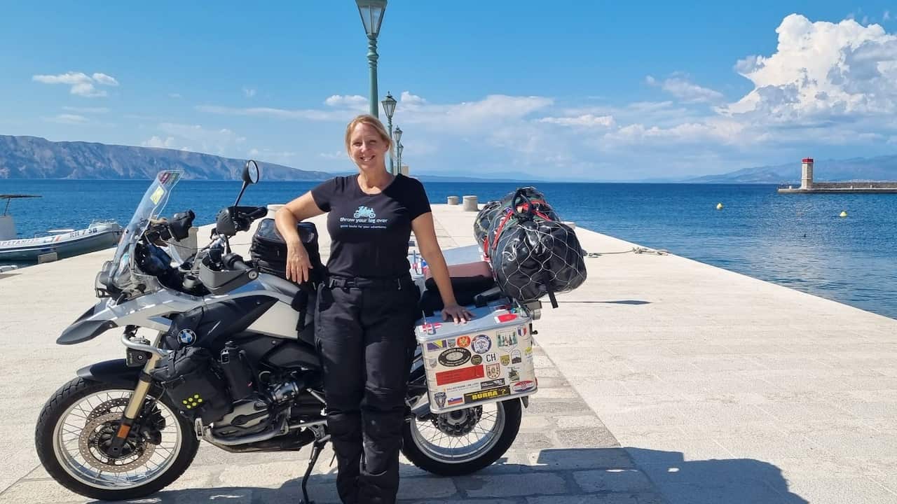 A middle-aged blonde woman stands in front of a motorcycle on a sandstone pier in front of water on a sunny day.