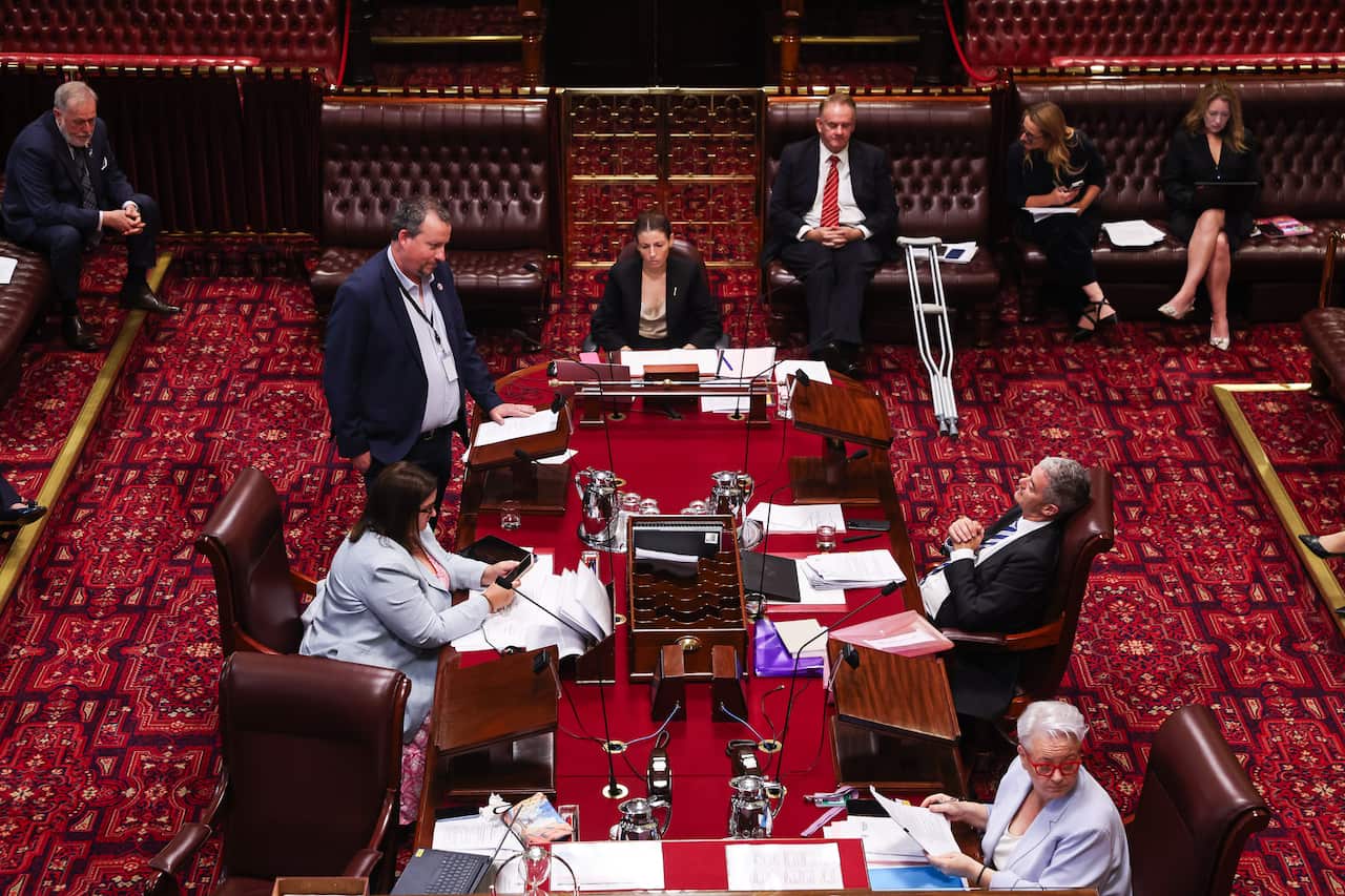 An overhead shot shows a group of people gathered around a long wooden table in a formal, red-carpeted chamber, likely a legislative assembly, with one man standing to address the others.