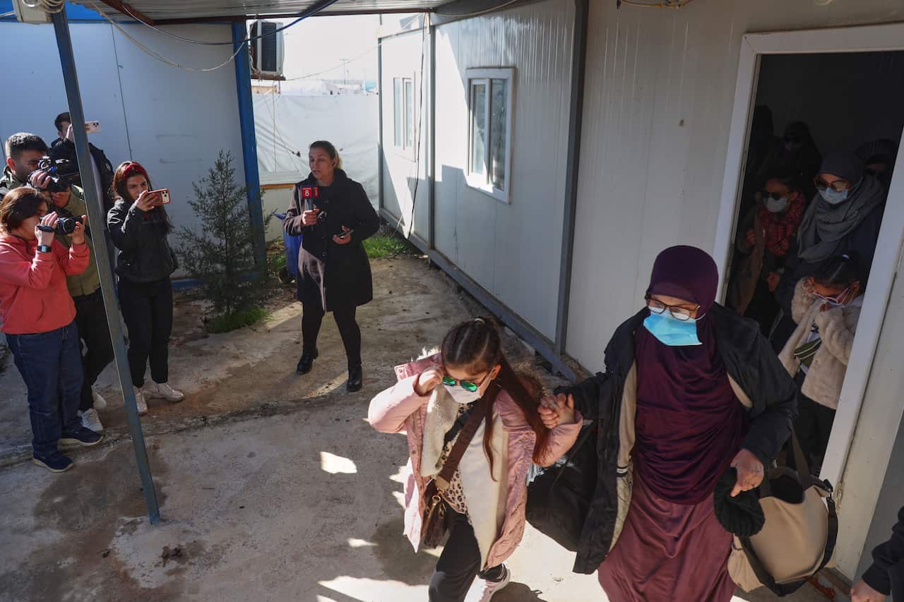 A group of Muslim women and children walking out of the door of a demountable. There is a small group of people opposite them filming with cameras and phones