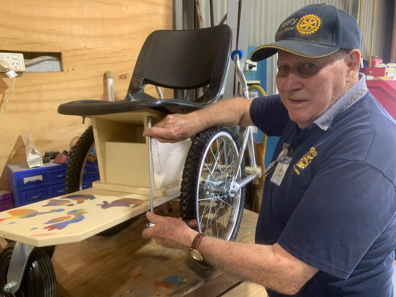 A man in a cap and blue t-shirt stands next to a wheelchair on a bench.