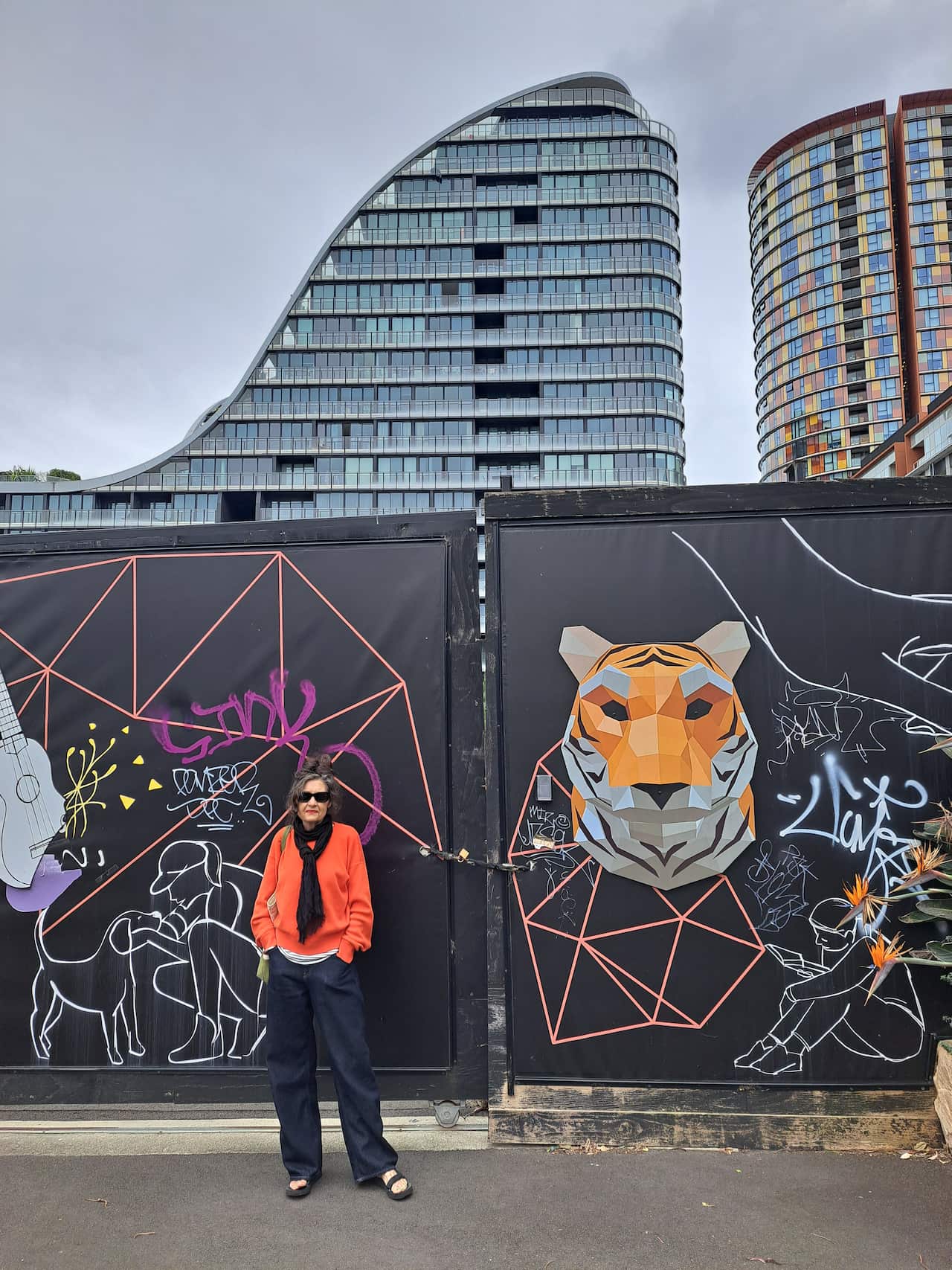 A woman wearing a bright red sweater stands in front of a large graffitied gate against a backdrop of high rise buildings.