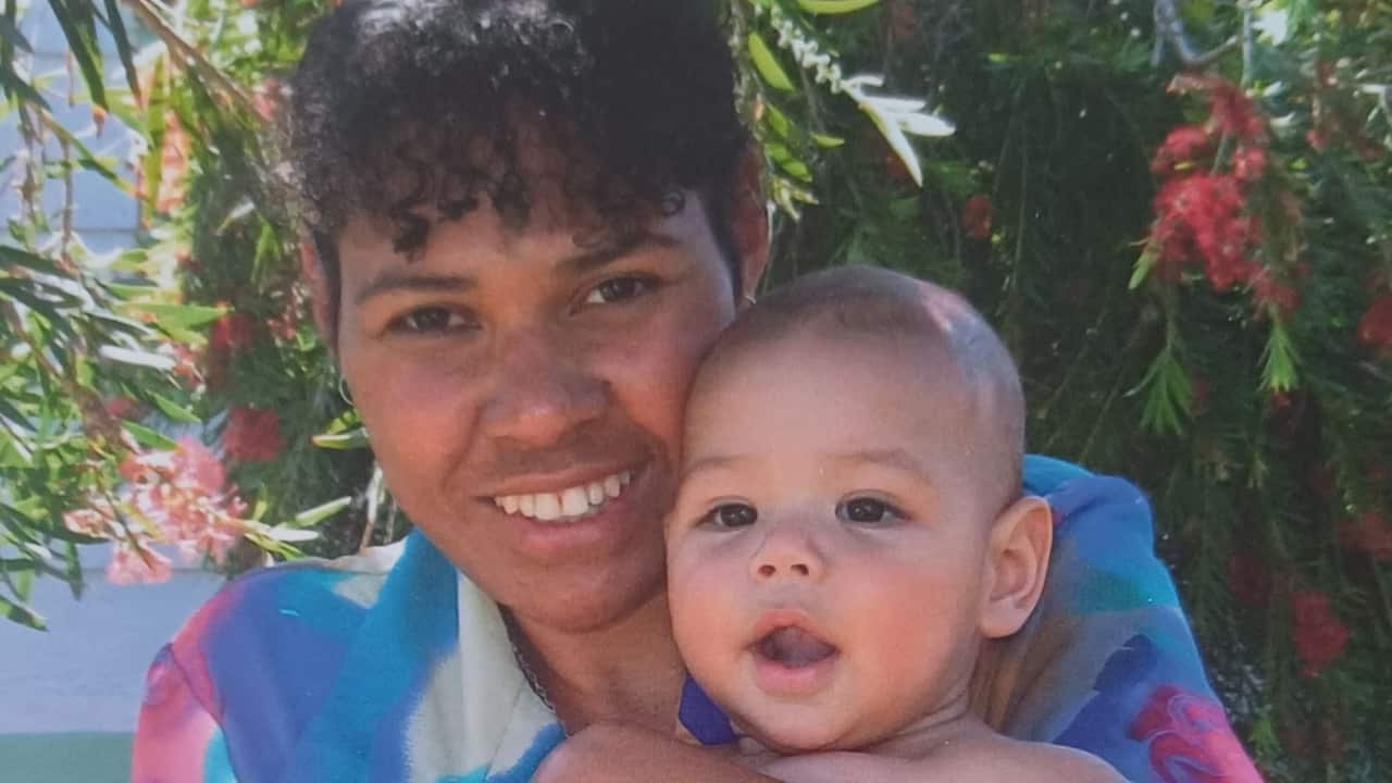 a young woman with dark curly hair smiles holding a young baby boy in front of green leaves in a backyard