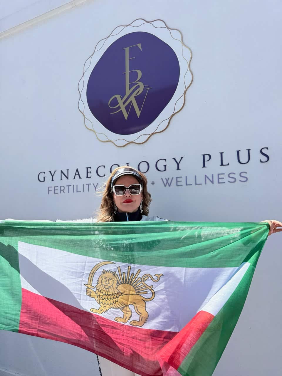 A woman wearing sunglasses stands in front of a clinic sign reading “Gynaecology Plus Fertility + Wellness,” holding an Iranian flag with a lion and sun emblem.
