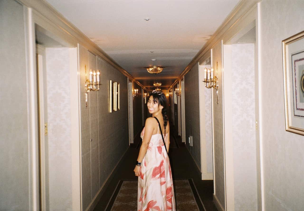 Girl in pink and white dress looks over her shoulder as she stands in a corridor.