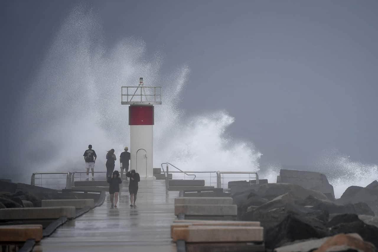Waves crash onto rocks as people look on the seaway.