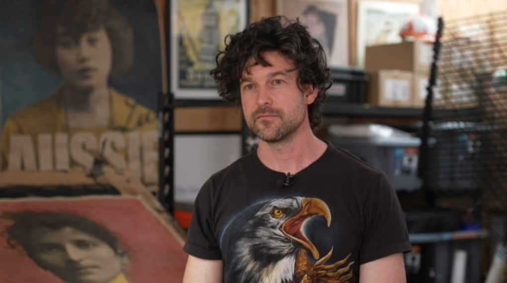 A man wearing a shirt with an eagle on it, with an art studio in the backdrop.