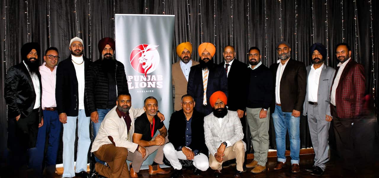A group of men from the Punjab Lions Adelaide club pose together for a group photo in front of a dark curtain backdrop and a branded banner.