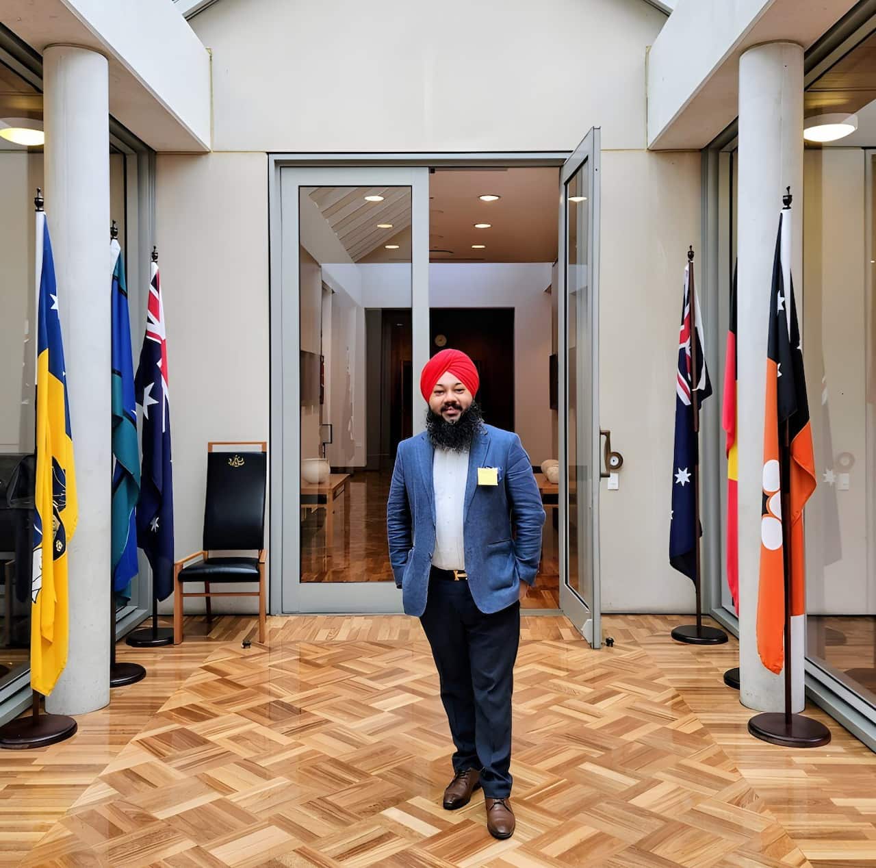 A man with a full beard wearing a red turban, a blue blazer, and dark pants stands in a hallway with a glass ceiling and parquet floor, flanked by columns displaying Australian and Aboriginal flags.