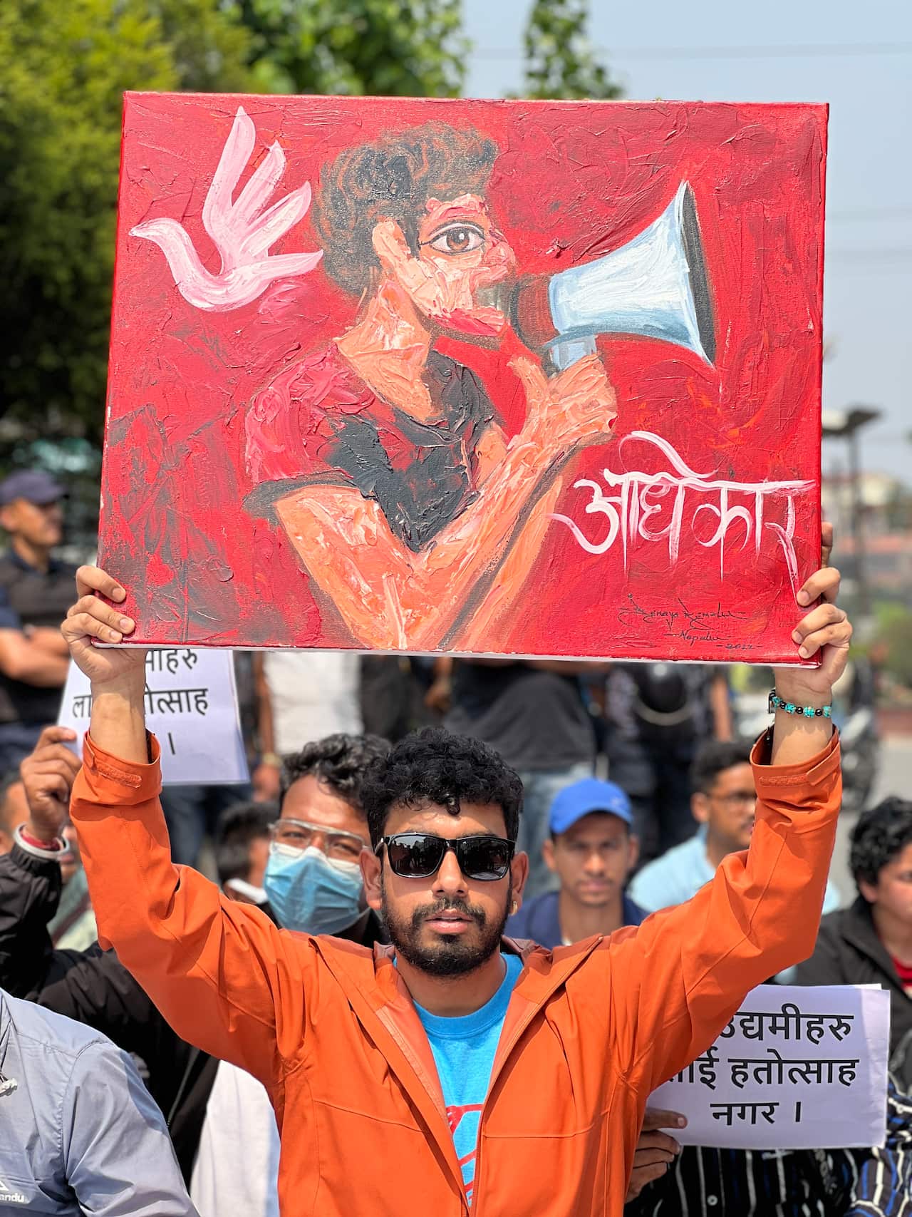 A man in an orange jacket holds up a painted sign at a protest.