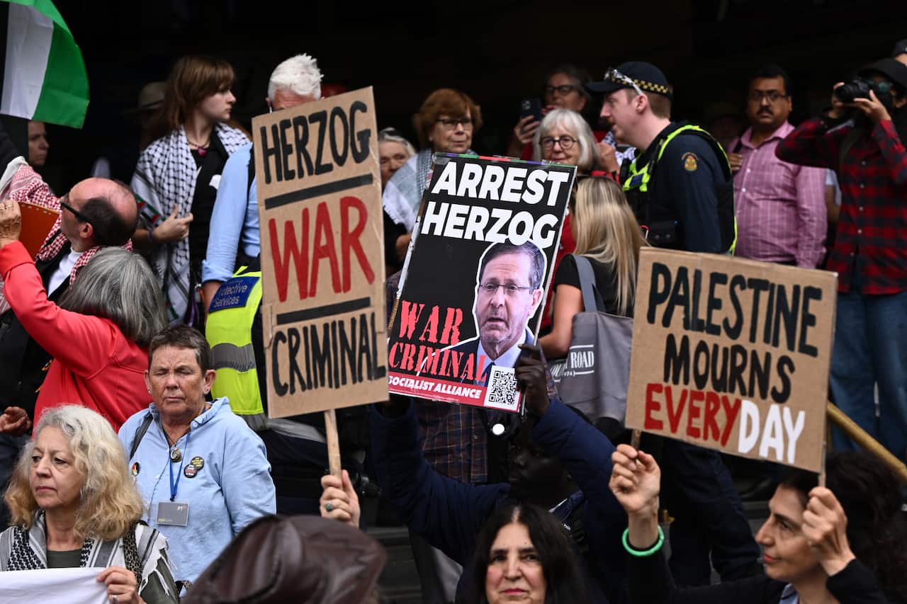 Protesters carrying signs accusing Israeli President Isaac Herzog of being a war criminal and calling for his arrest. 