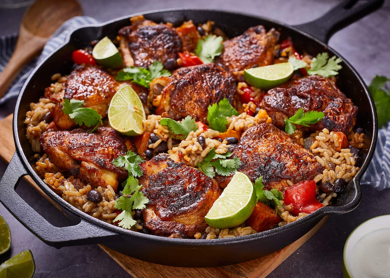 A round black cast-iron baking dish holds cooked chicken and rice. The dish is garnished with fresh herbs and lime wedges. A striped cloth and wooden spoon can be seen sitting behind the dish on one side.