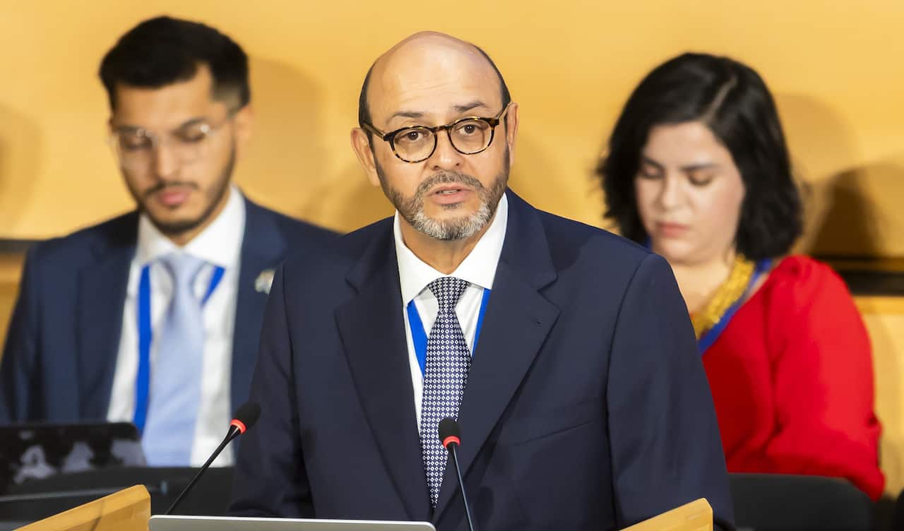 A man speaking at a podium. A man and a woman are behind him.
