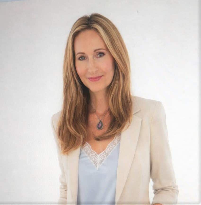 A woman with long blonde hair wearing a beige jacket and light blue top faces camera.