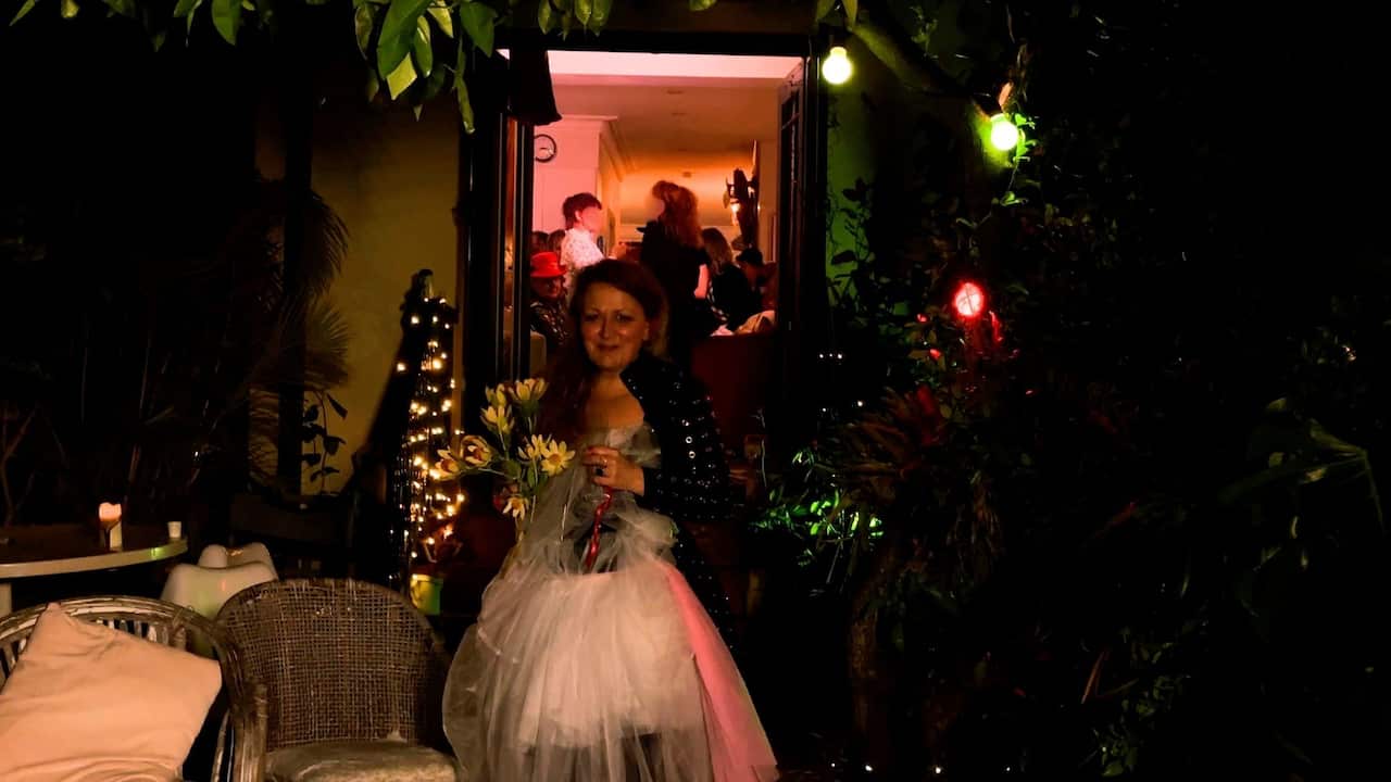 a middle aged woman in a tutu holds flowers and stands in a backyard that is dimly lit