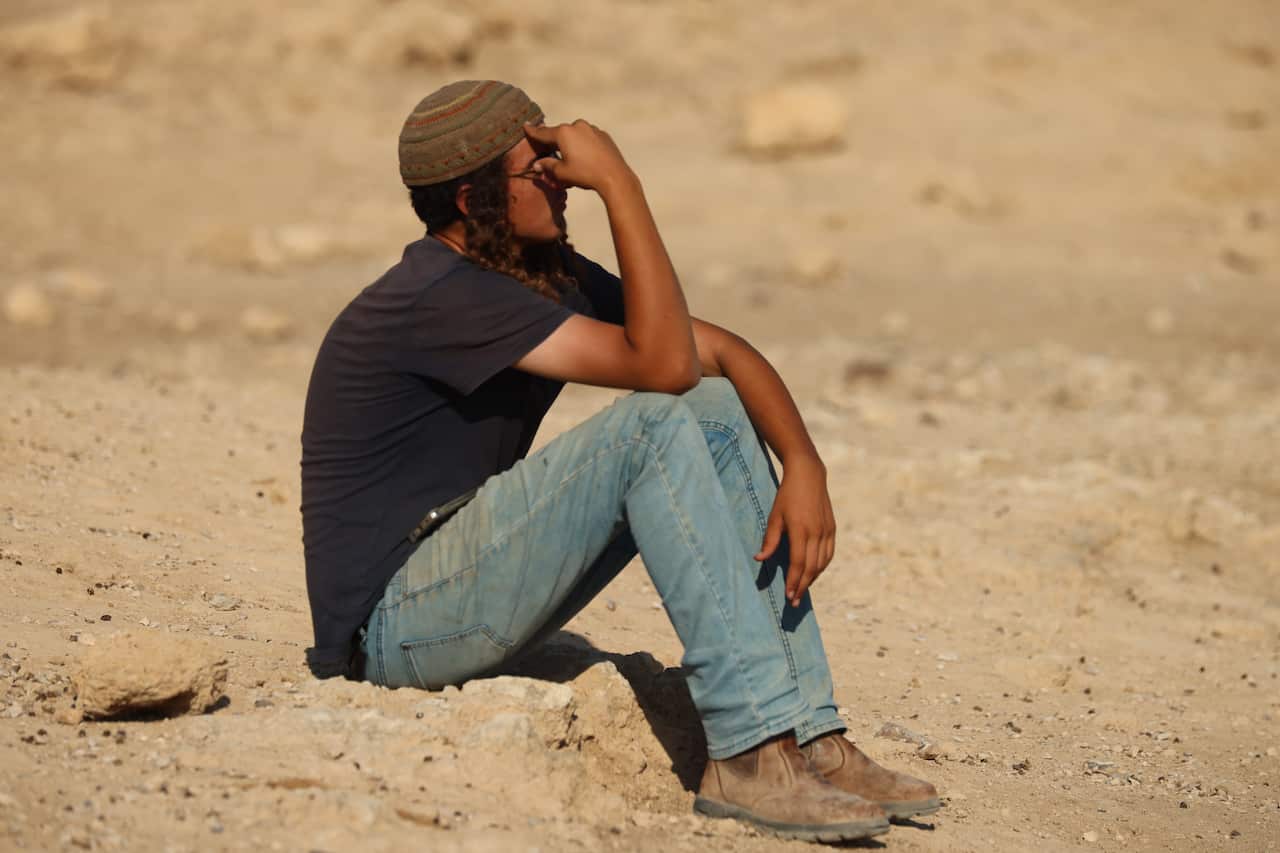 A teenage boy wearing blue jeans, a black t-shirt and a kippah sits on rocks with a hand over his face. 
