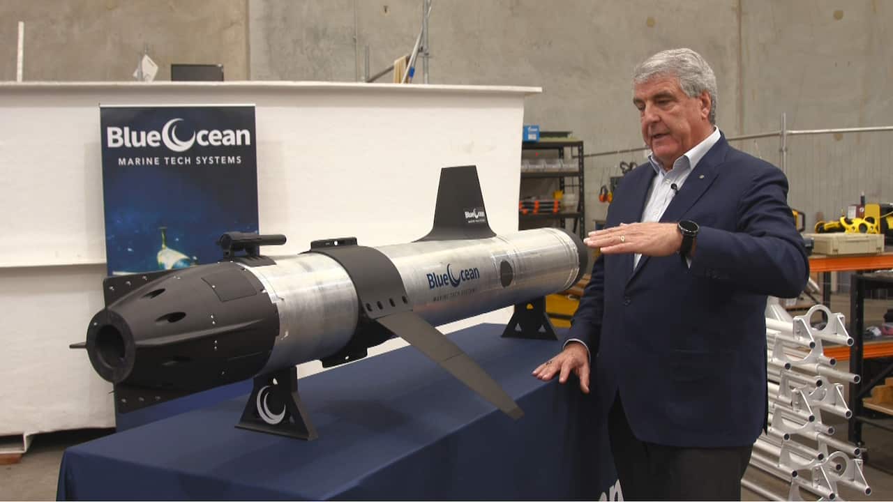 Middle aged man demonstrating a underwater drone vehicle next to him on display on a blue table.