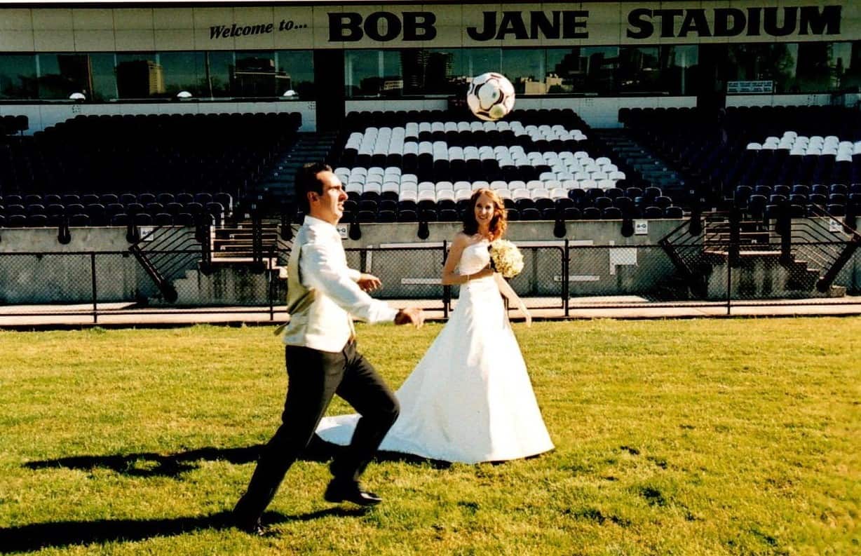 A man wearing black pants and a white shirt juggles a football on a football pitch in front of his wife, who is wearing a wedding gown.