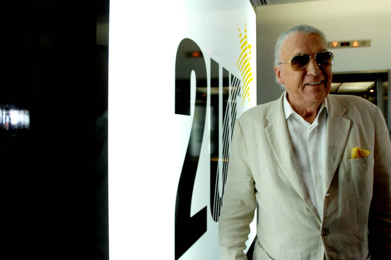 An older man wearing a cream suit and sunglasses smiles as he walks past a large white sign with 2UE written on it in large white letters