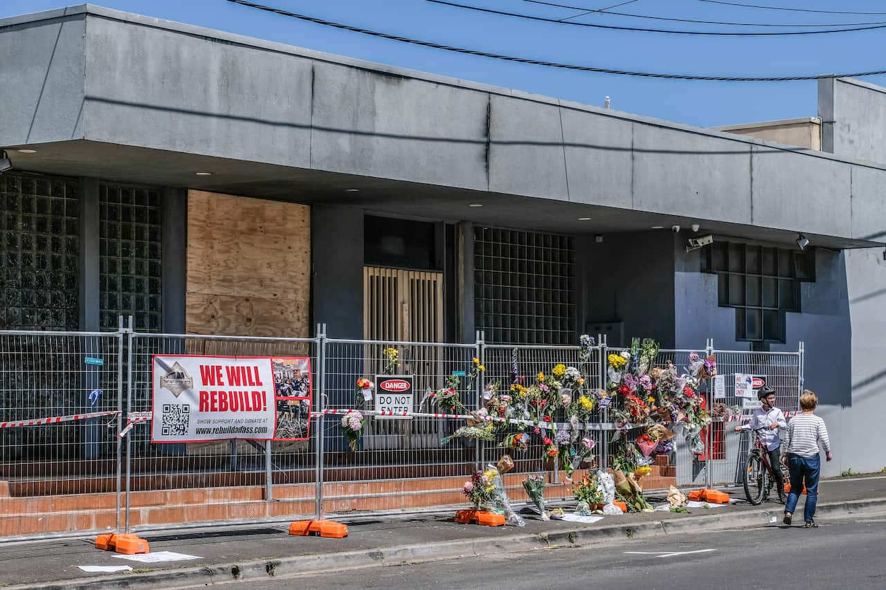 A fence with flowers stuck to it, and a one storey building behind it that is damaged.