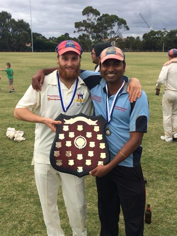 Two cricket players wearing medals stand together on a grassy field, smiling as they jointly hold up a dark wooden championship shield adorned with small silver plaques.