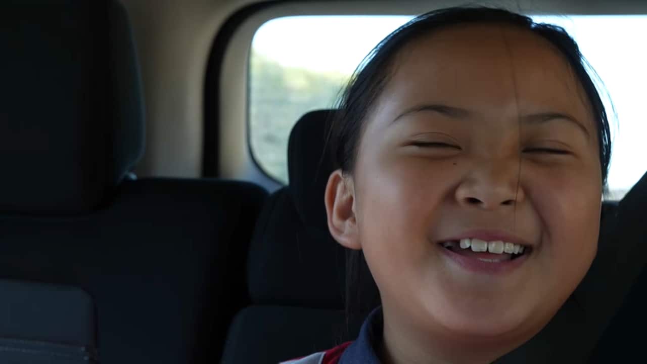 A smiling young girl sits in the back seat of a car, looking toward the camera.