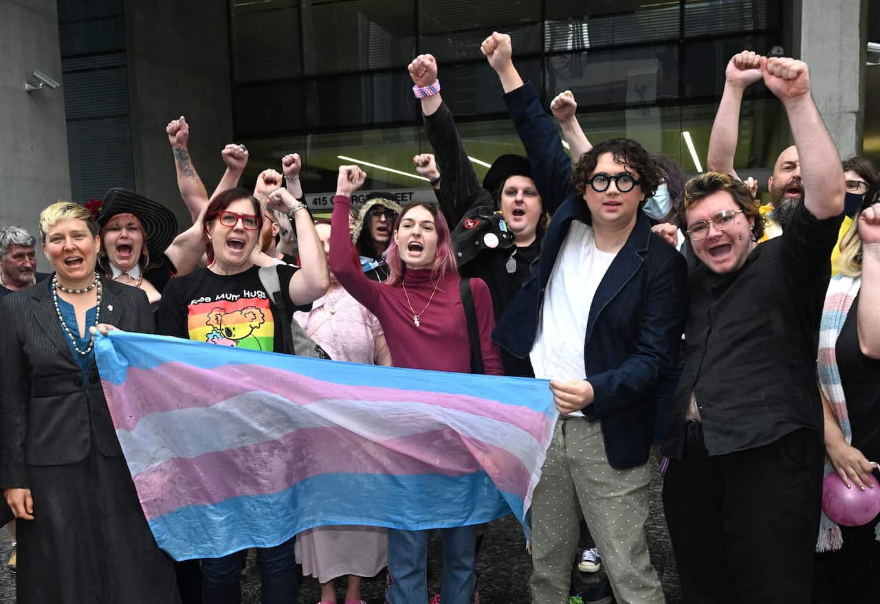 A group of transgender advocates celebrating outside court. Most have their hands raised in the air. A transgender flag is being held.
