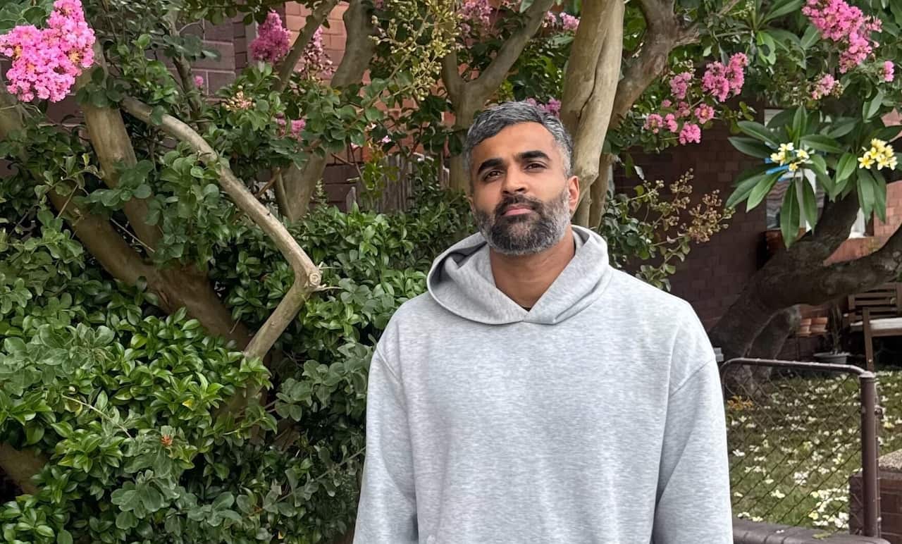 A man wearing a grey hoodie, standing in front of trees.