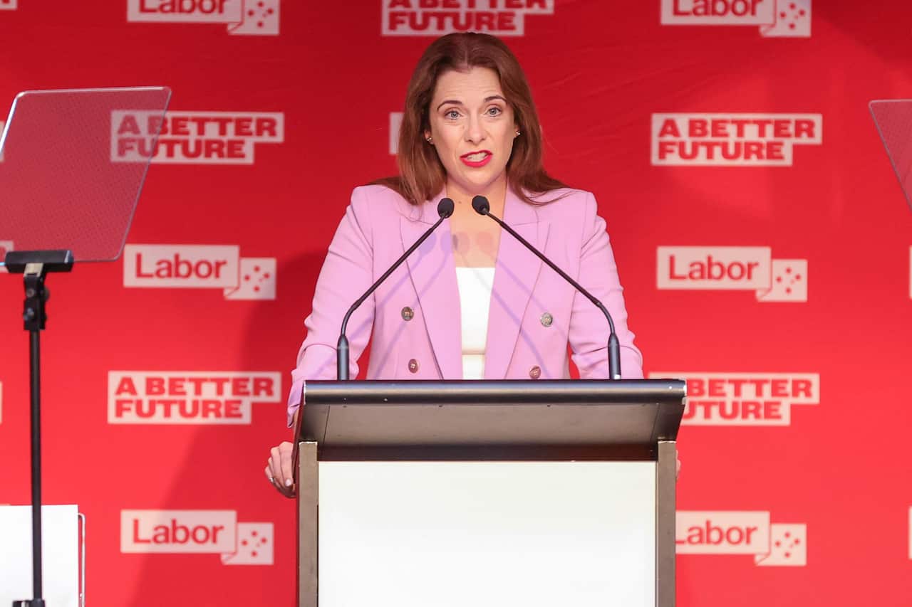 A woman with red hair stands at a lectern with Labor campaign signage behind her