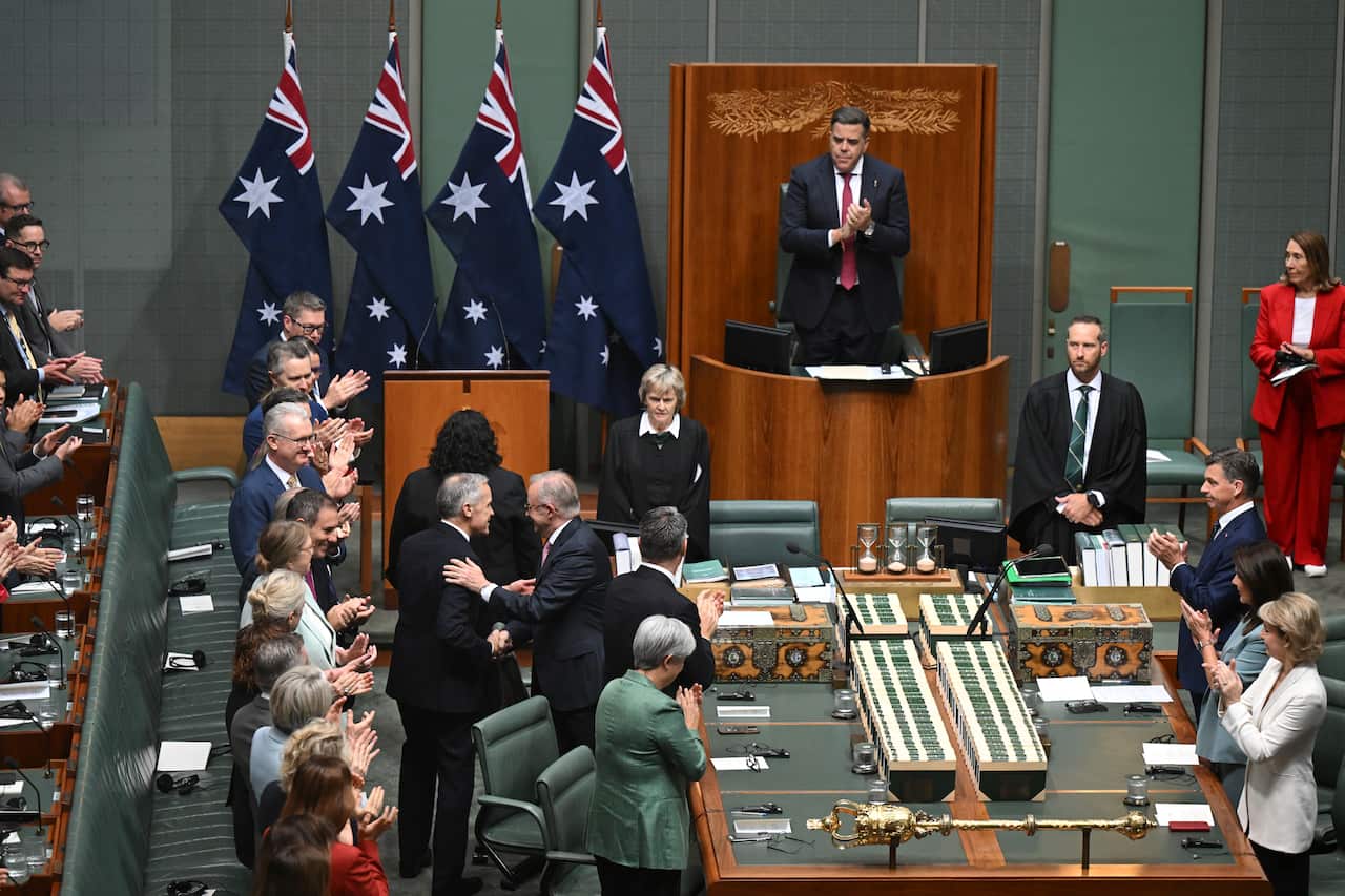 Mark Carney and Anthony Albanese shaking hands in a large room with green walls and a row of Australian flags. Several people are applauding.